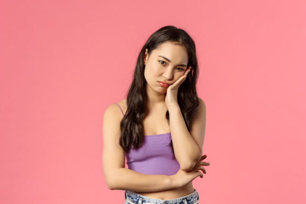 A young woman standing and looking bored staring ahead bored, pink background