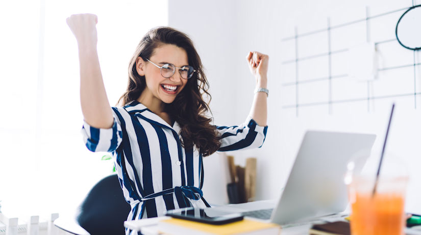 Excited young woman sitting at table with laptop and celebrating success